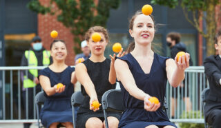 Three jugglers dressed in smart navy dresses sit in a row on black fold out chairs. They are each juggling 4 oranges.