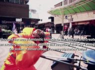 A street performer in an orange outfit entertains a crowd in a city square, with shops and people in the background. Overlaid text quotes Zoe Stokes about the excitement of the Coventry Mysteries Festival.