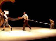 Three performers are on stage; one pulls a long strap tied around the waists of the other two, who resist being pulled forward. The background is dark, with a large, curved wooden structure on the left.