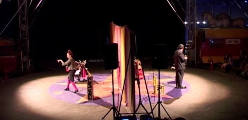 A circus ring with two performers stands in the spotlight; one is in a colorful costume on the left, gesturing animatedly, while the other stands on the right in a suit. The audience and circus decor are dimly visible in the background.