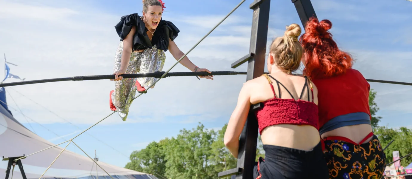 A circus performer in a sparkly costume balances on a high wire, smiling at two women supporting the wire. A circus tent and festival attendees are visible in the background on a sunny day.
