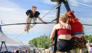 A circus performer in a sparkly costume balances on a high wire, smiling at two women supporting the wire. A circus tent and festival attendees are visible in the background on a sunny day.