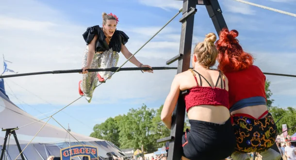 A circus performer in a sparkly costume balances on a high wire, smiling at two women supporting the wire. A circus tent and festival attendees are visible in the background on a sunny day.