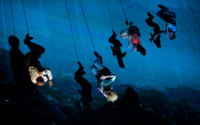 Four people appear to be walking or climbing on a vertical blue surface, with dramatic shadows cast behind them, creating an illusion of defying gravity.