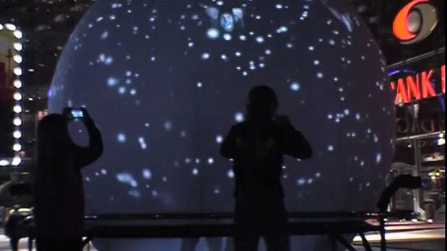 Two people are silhouetted in front of a large, spherical display with white, star-like dots, while city lights and signs are visible in the background.