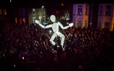 A large crowd gathers at night around a tall, illuminated humanoid puppet with outstretched arms, set against the backdrop of residential houses and lit street lamps.