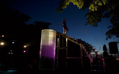 A person stands atop a tall, illuminated structure with stairs and a transparent cylinder, silhouetted against a deep blue evening sky, with trees and stage lights in the background.