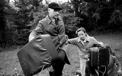 A man and a woman carrying suitcases climb up a grassy hill, dressed in old-fashioned coats. The man wears a flat cap and leads, while the woman follows closely behind him. Trees fill the background.