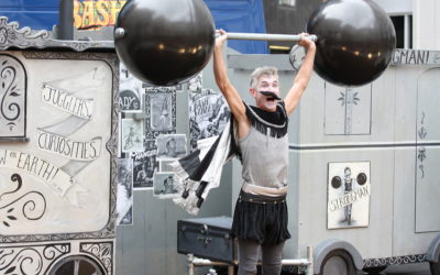 A street performer dressed as an old-time strongman lifts a large, fake barbell overhead. He wears a fake mustache, face paint, and vintage circus-style clothing, performing in front of decorated circus-themed props.