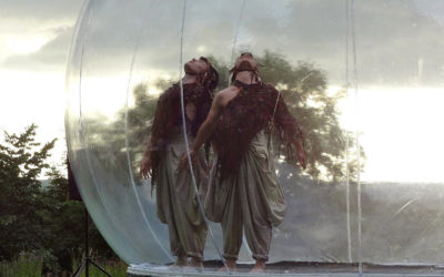 Two dancers dressed in loose pants and feathered tops stand back to back inside a large transparent inflatable sphere, looking upward, with trees and a cloudy sky visible in the background.