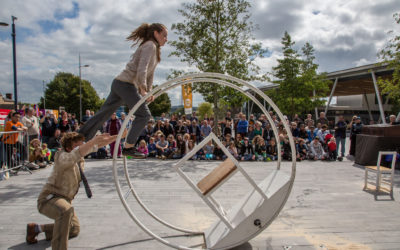 A performer balances on a large metal wheel while another pushes it; a crowd watches the outdoor street performance on a cloudy day, with trees and buildings in the background.