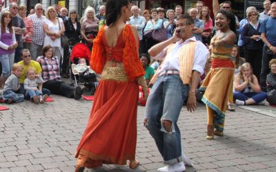 Three street performers in colorful costumes dance energetically on a cobblestone street, surrounded by a large, engaged crowd of onlookers of various ages.