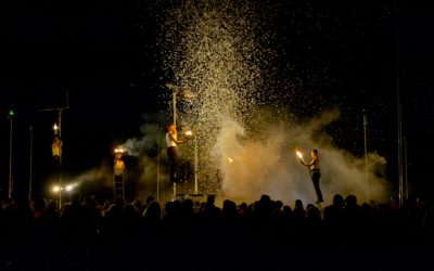 Performers juggle fire on stage at night with artificial snow falling and smoke around them, while a crowd watches in the foreground. Some performers are elevated on platforms, creating a dramatic scene.