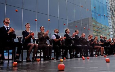 A group of people in formal attire sit in a row on chairs, simultaneously juggling apples in front of a modern glass building, with several apples on the ground.