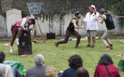 Four performers act out a lively outdoor scene in front of an audience, with one person climbing a block, two interacting animatedly, and one watching. A Caution Land Mine sign is in the background near a brick wall.