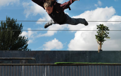 A person with long hair is captured mid-air, arms and legs outstretched, appearing to jump or fall above a rooftop with a small potted tree and a blue sky with clouds in the background.