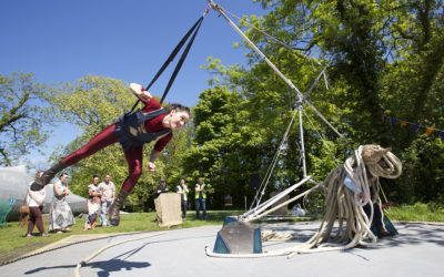 A person in a harness is swinging from a metal structure outdoors, surrounded by trees and grass. Several people watch nearby, and thick ropes are coiled on the ground next to the equipment.