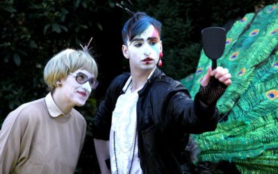 Two people in costume and dramatic makeup stand outdoors. One wears glasses and a beige sweater, the other has colorful makeup, a black jacket, and holds a hand mirror. Large green peacock feathers are attached behind them.