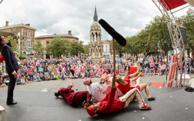 A lively outdoor street performance shows a crowd watching actors in colorful costumes lying on stage, while a suited performer stands to the side. Historic buildings and a stone monument are visible in the background.