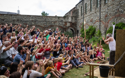A large crowd, including many children, sits outdoors near a stone building, eagerly raising their hands toward a performer on a small stage who is engaging with them energetically.