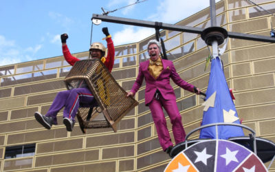 Two performers are elevated in the air; one sits in a metal basket, smiling and wearing a helmet, while the other stands beside him in a purple suit, holding onto a pole. Both appear joyful, with a building in the background.