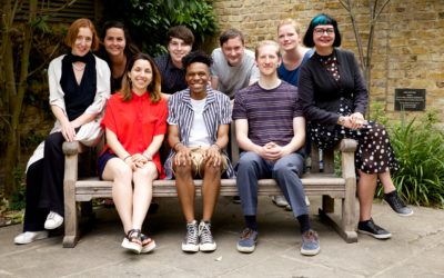A group of ten people, with varied appearances and styles, pose together outdoors on and around a wooden bench in front of a brick wall and greenery, smiling warmly at the camera.