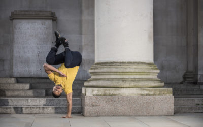 A man in a yellow shirt and black pants performs a one-handed handstand against a column, with stone steps and an engraved plaque in the background.