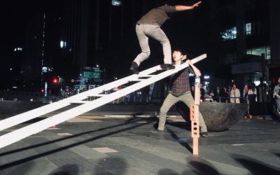A person balances on an angled wooden beam while another person stands below, ready to assist, during a nighttime outdoor performance with an audience watching in the background.