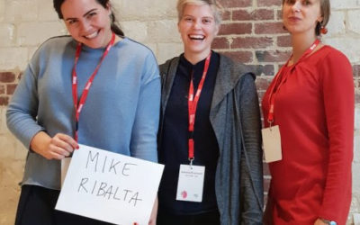 Three women wearing lanyards stand in front of a brick wall. The woman on the left is smiling and holding a sign that reads MIKE RIBALTA. The other two women are also smiling and standing close together.