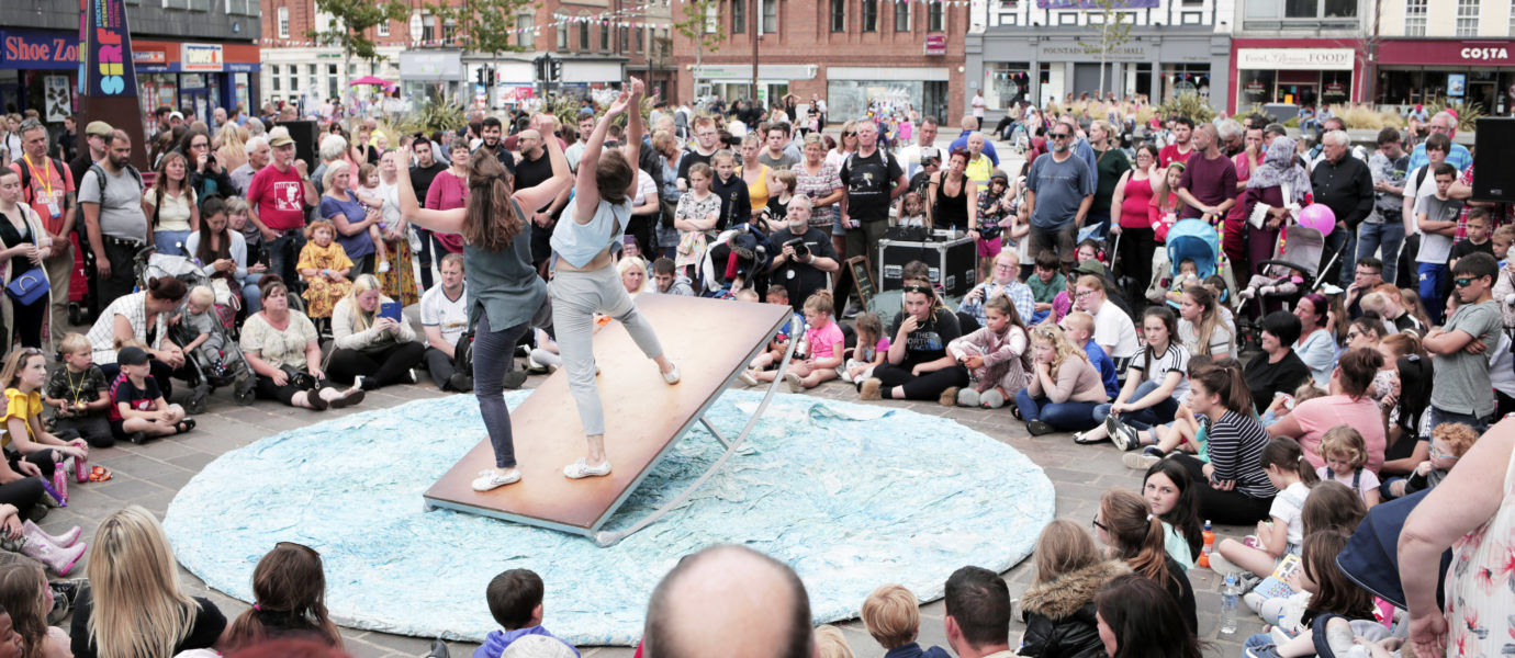Two performers balance on a tilted platform in the center of a circular stage, surrounded by a large crowd of people watching outdoors in an urban plaza.