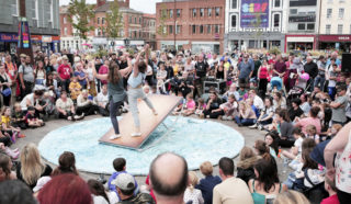 Two performers balance on a tilted platform in the center of a circular stage, surrounded by a large crowd of people watching outdoors in an urban plaza.
