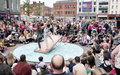 Two performers balance on a tilted platform in the center of a circular stage, surrounded by a large crowd of people watching outdoors in an urban plaza.