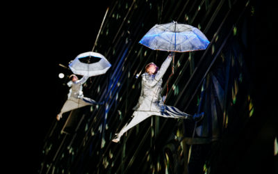 Two performers in silver costumes dangle from harnesses on a building at night, each holding a glowing umbrella, creating a whimsical, gravity-defying scene with light patterns on the wall behind them.