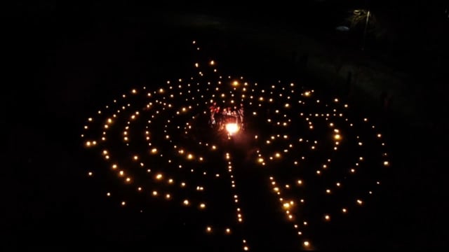 Aerial view of a labyrinth pattern formed by small lights in the dark, with a group of people gathered around a bright central light or fire at the center.