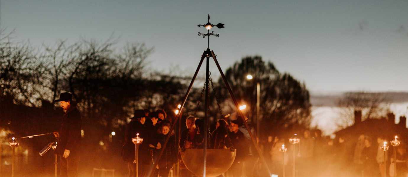 A group of people gather outdoors at dusk around a large suspended cauldron, surrounded by torches and glowing lights arranged on the grass, creating a warm, atmospheric scene.
