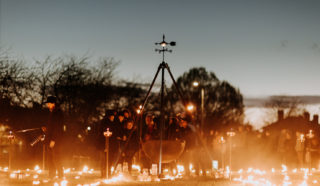 A group of people gather outdoors at dusk around a large suspended cauldron, surrounded by torches and glowing lights arranged on the grass, creating a warm, atmospheric scene.