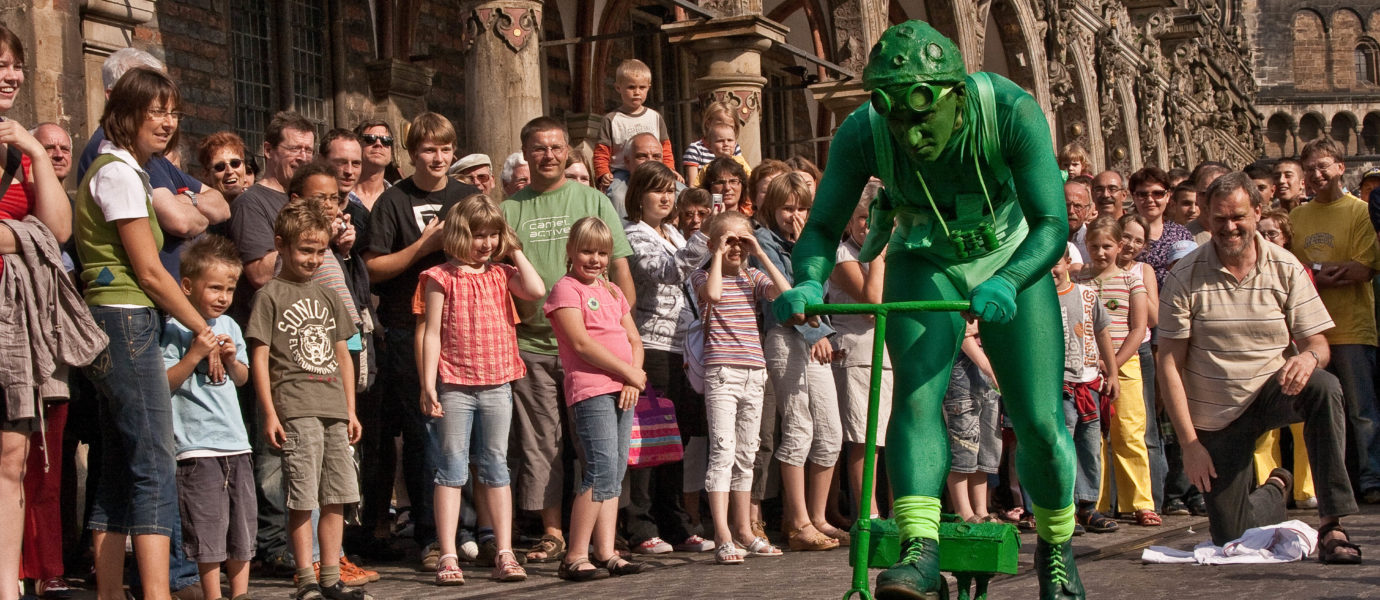 A performer dressed in a green frog costume rides a small green scooter along a cobblestone street, entertaining a large crowd of people standing and watching outside historic buildings.