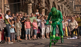 A performer dressed in a green frog costume rides a small green scooter along a cobblestone street, entertaining a large crowd of people standing and watching outside historic buildings.
