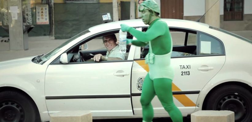 A man dressed in a green superhero costume walks past a white taxi, while the taxi driver smiles and looks at him through the open window. The scene appears to be in an urban area during the day.