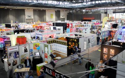 Large convention hall filled with numerous colorful booths, banners, and displays, with many people walking, browsing, and interacting at a busy trade show or expo event. Bright lighting and a lively atmosphere.