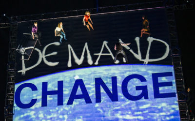 Performers are suspended in mid-air against a large backdrop reading DEMAND CHANGE in bold white and blue letters, with a dark background and stage lighting highlighting the scene.
