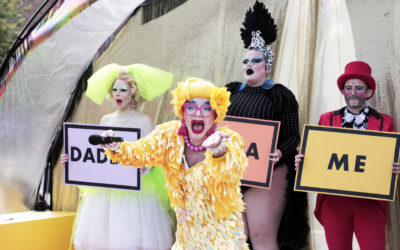 Four drag performers are on stage. The center performer, in a yellow feathered costume and bright wig, holds a microphone and reaches out. The others hold signs reading DADDY, A, and ME against a gold backdrop.