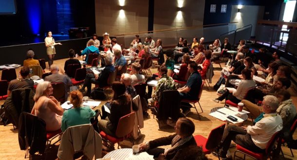 A group of people sit on red chairs arranged in small circles in a spacious room, listening to a woman speaking at the front. The room has wooden floors, high ceilings, and spotlights illuminating the scene.