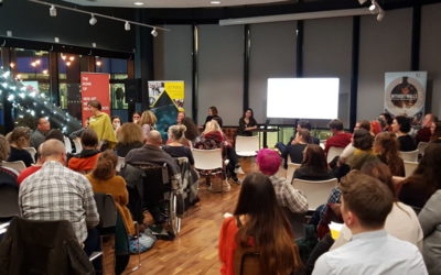 A diverse group of people sits facing a panel of speakers in a modern, well-lit room with large windows, string lights, and a projection screen at the front. Some audience members are engaged in conversation.