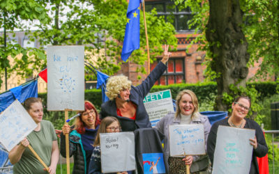 Five people stand outdoors holding protest signs supporting womens rights and the EU. One person in a blond wig makes a peace sign. EU flags and trees are in the background. The mood is positive and cheerful.
