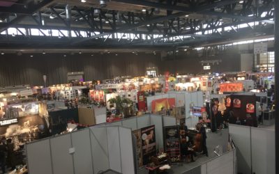 A large indoor convention hall filled with numerous booths, colorful displays, posters, and people walking around and interacting, under a high ceiling with exposed beams and lights.