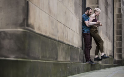 Two people stand in a doorway on a stone building; one person leans against the frame while the other steps carefully down to the pavement. Both seem focused and casual, with a soft, urban background.