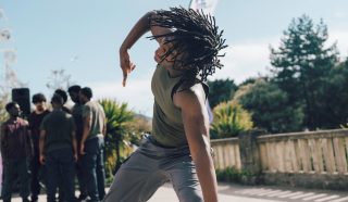 A person with dreadlocks performs an expressive dance move outdoors, wearing a green sleeveless shirt and gray pants. A group of people stands in the background near greenery and a stone railing.