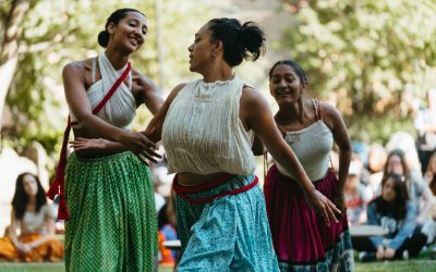 Three women in colourful skirts and sleeveless tops dance gracefully outdoors, surrounded by a blurred audience and trees in the background, enjoying the lively atmosphere.