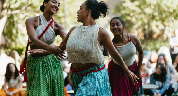 Three women in colourful skirts and sleeveless tops dance gracefully outdoors, surrounded by a blurred audience and trees in the background, enjoying the lively atmosphere.